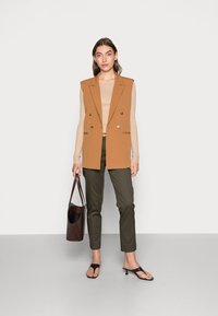 Bruine mouwloze blazer, beige geribbelde top, olijfgroene broek, donkerbruine tas en zwarte hakken. Soepele texturen en een nauwsluitende pasvorm.