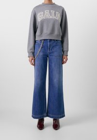 Grijze sweatshirt met de tekst "GAIA" in het wit, gecombineerd met blauwe wide-leg jeans met een kettingdetail op de tailleband en bordeaux schoenen.