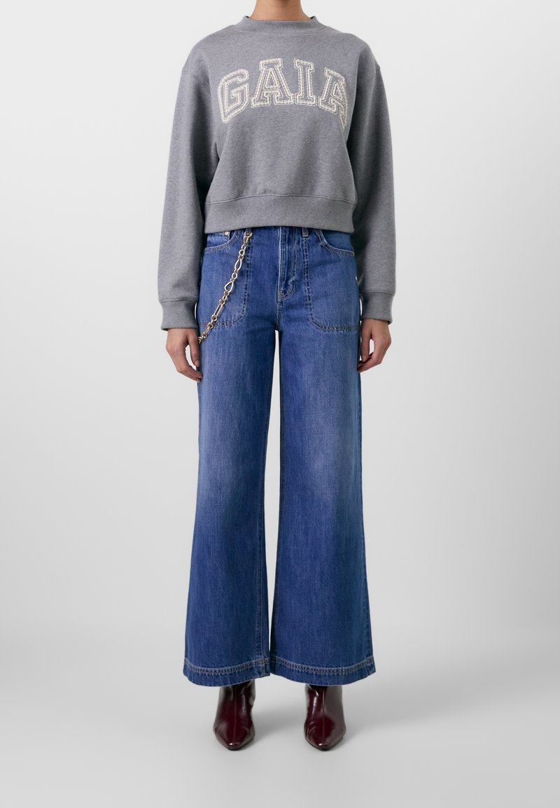 Grijze sweatshirt met de tekst "GAIA" in het wit, gecombineerd met blauwe wide-leg jeans met een kettingdetail op de tailleband en bordeaux schoenen.