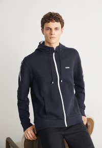 Marinblå zip-up hoodies i bomullsblandning med vit dragkedja och logotyp. Har en huva, sidofickor och kontrasterande vita ränder på ärmarna.