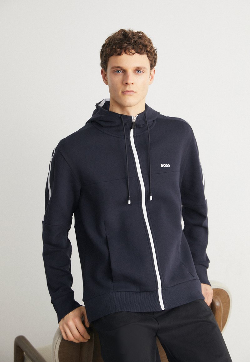 Marinblå zip-up hoodies i bomullsblandning med vit dragkedja och logotyp. Har en huva, sidofickor och kontrasterande vita ränder på ärmarna.