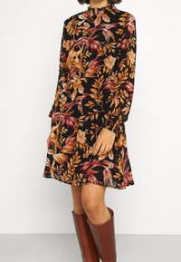 Kukkamekko mustana oranssi- ja punakukkaisten kuvioiden kanssa. Varustettu pitkillä hihoilla, rypytetyllä kauluksella ja rentoa istuvuutta. Yhdistetty ruskeisiin polvipitkiin saappaisiin.