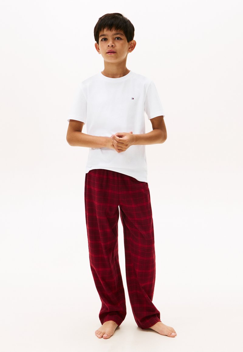 Garçon debout pieds nus portant un t-shirt blanc à manches courtes et un pantalon de pyjama ample à carreaux rouges sur fond blanc.