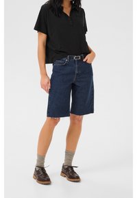 Mörkblå denimshorts med fransig nederkant, matchat med en svart pikétröja med kort ärm. Bruna skor med tjock sula och grå strumpor synliga.