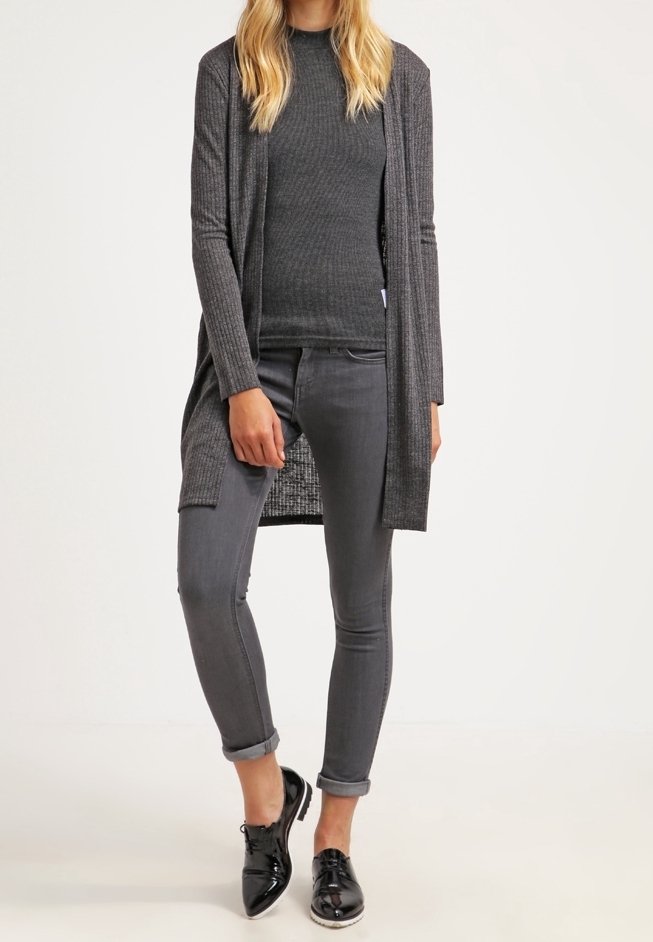 Un long cardigan gris sur un haut à côtes gris, associé à un jean gris retroussé aux chevilles et des chaussures en cuir verni noires.