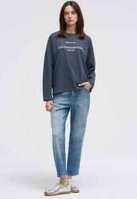 Dunkelgrauer langärmeliger Sweatshirt mit weißem Text, kombiniert mit hellblauen geraden Jeans und metallisch silbernen Sneakers. Einfaches Design.