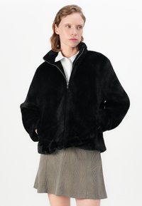 Zwarte nepbont zip-up jack met een hoge kraag, oversized pasvorm en twee zijzakken. Draagt over een geruite rok.