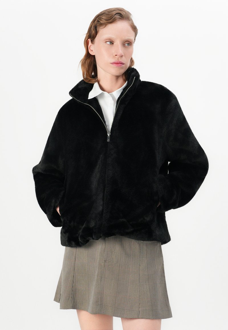 Veste zippée en fausse fourrure noire avec un col montant, coupe oversize et deux poches latérales. Portée sur une jupe à carreaux.