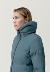 Vadderad jacka i teal-färg, med hög krage, dragkedja fram och dekorativ sömnad på ärmarna med små accentdetaljer.