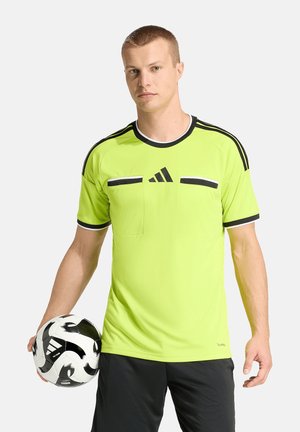 Junger Mann trägt ein neongelbes Sporttrikot mit schwarzen Streifen und hält einen schwarz-weißen Adidas-Fußball in der linken Hand.