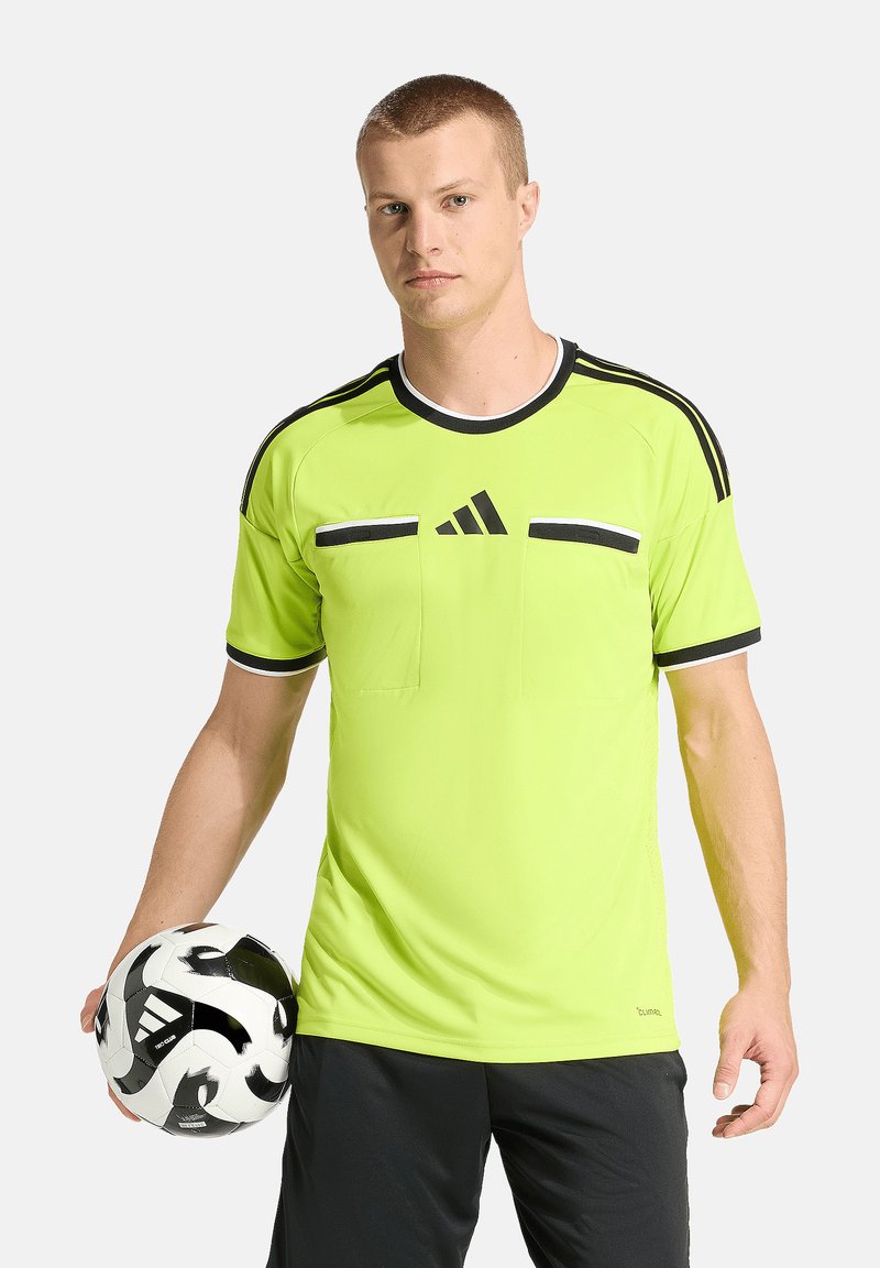 Jeune homme portant un maillot de sport jaune néon avec des rayures noires, tenant un ballon de football Adidas noir et blanc dans sa main gauche.