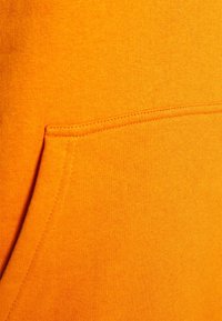 Tejido de sudadera naranja con una textura suave. Presenta un bolsillo tipo canguro cosido con hilo a juego, que enfatiza un diseño limpio.