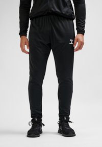 Mężczyzna w czarnych sportowych spodniach dresowych i czarnych butach sportowych, stojący na tle jednolitego jasnego tła.