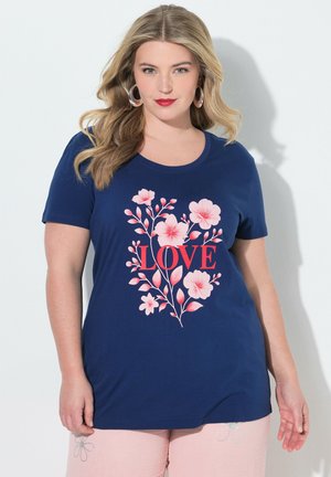 Kvinde iført en mørkeblå T-shirt med lyserødt blomstermotiv og ordet "LOVE" i rødt, kombineret med lyserøde bukser.