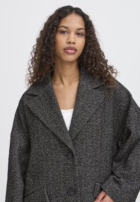 Ung kvinde med krøllet hår iført en mørkegrå hørmønsteret frakke med store revers og sorte knapper over en hvid bluse.