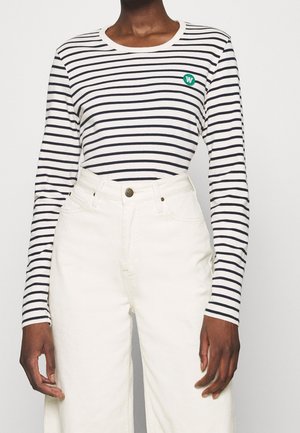 Henkilö, jolla on valkoinen pitkähihainen paita, jossa on mustia vaakaraitoja ja pieni vihreä logo, yhdistettynä korkeavyötäröisiin off-white-housuihin.