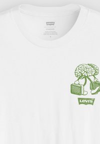 Balta kokvilnas T-kreklā ar zaļu grafiku, kas attēlo smadzenes ar kājām, kas nes portfeli, un apakšā ir "Levi's" logo.