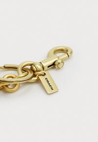 Gold-toned metal clasp with a rounded hook, interlocking rings, and a tag inscribed with "COACH," featuring a smooth finish and polished texture.