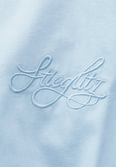 Ανοιχτό μπλε ύφασμα με λευκή κεντημένη καλλιγραφική λέξη «Lacedlit» στο κέντρο του υλικού.