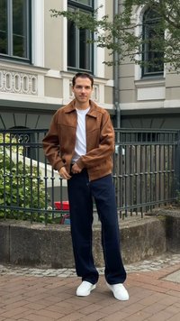 Giaccavento in suede marrone con tasche con zip, t-shirt bianca, pantaloni a gamba larga blu scuro e sneakers bianche, sullo sfondo di un edificio.