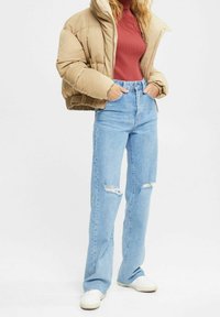 Chaqueta acolchada beige, diseño corto, suéter rojo acanalado, jeans azul claro de pierna recta con detalles desgastados y zapatillas blancas.