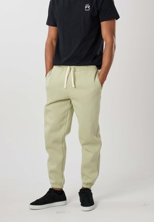 Mężczyzna w beżowych spodniach dresowych z białym sznurkiem, czarnych tenisówkach i czarnej koszulce z małym białym nadrukiem na piersi. Ręce w kieszeniach.