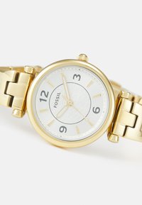 Goldfarbene Fossil-Uhr mit rundem Gehäuse, weißem Zifferblatt, erhöhten Stundenmarkierungen und strukturiertem Zentrum. Metallarmband mit Druckknopfverschluss.