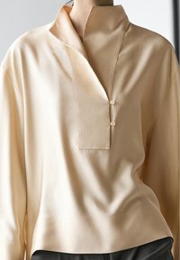 Beige blouse met lange mouwen, een hoge kraag en twee knopen aan de voorkant, gedragen over donkergrijze broek.