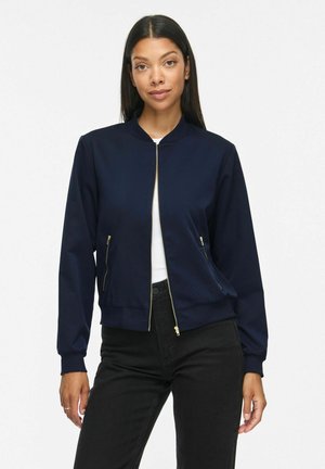 Femme portant une veste bomber bleu marine avec des fermetures éclair dorées sur un haut blanc et un pantalon noir, debout devant un fond blanc uni.