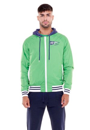 Un homme porte une veste verte à capuche et à fermeture éclair, avec des finitions bleu marine, ainsi qu’un pantalon bleu marine assorti, sur fond blanc.