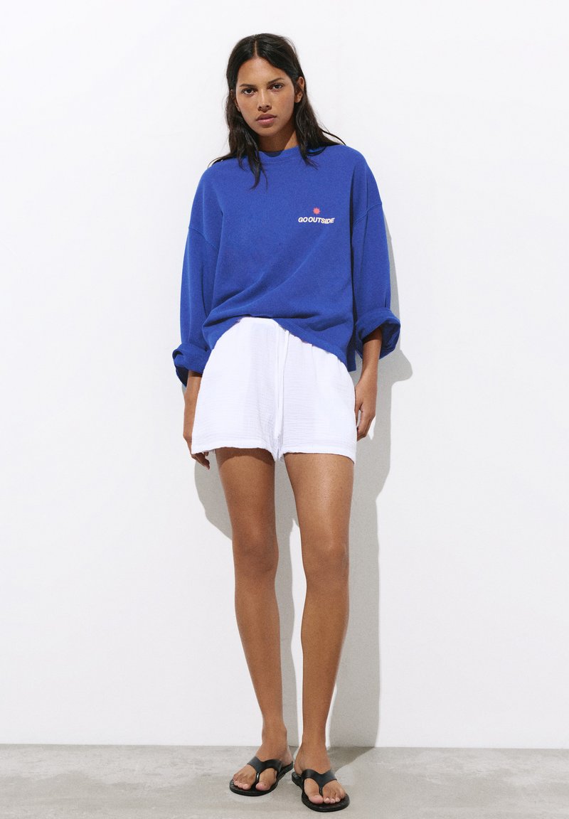 Femme portant un t-shirt bleu à manches longues avec le texte « GO OUTSIDE », un short blanc et des sandales noires, debout contre un mur blanc.