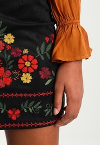 Schwarzer Rock mit buntem floralen Stickerei in Rot, Gelb und Grün. Verfügt über eine rüschige orangefarbene Bluse mit langen Ärmeln. Strukturiertes, geschichtetes Design.