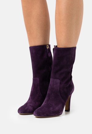 Fialové semišové kotníkové boty s špičatou špičkou a tenkým vysokým podpatkem. Mají boční zip a hladkou texturu.