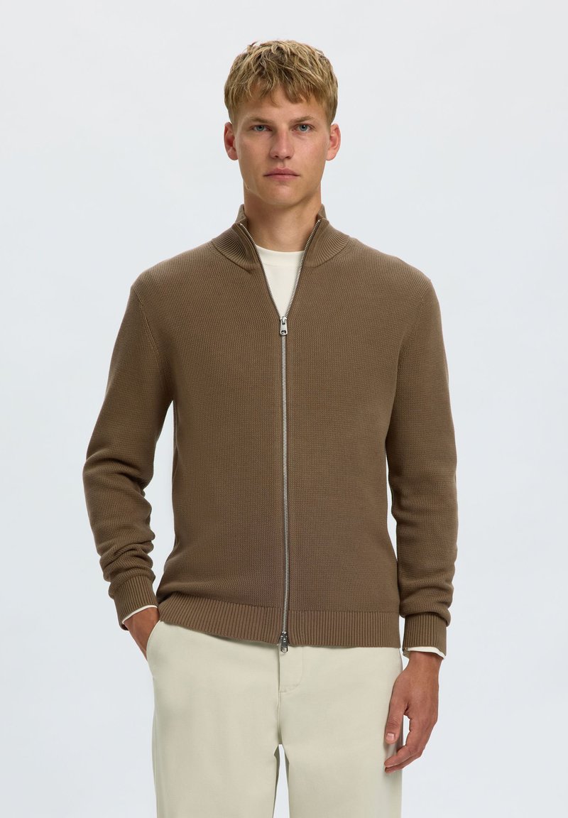 Jeune homme aux cheveux blonds courts, portant un pull marron zippé et un pantalon crème, debout devant un fond clair uni.