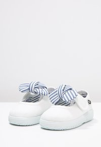Zapatillas de lona blancas con suela de goma, que presentan un lazo a rayas azul y blanco en la parte delantera y una correa de velcro para el cierre.