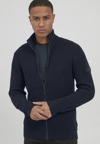 Maglione lavorato a maglia di un blu navy scuro con colletto alto, chiusura con zip, polsini a coste e un tessuto testurizzato. Presenta una patch con logo sul braccio.