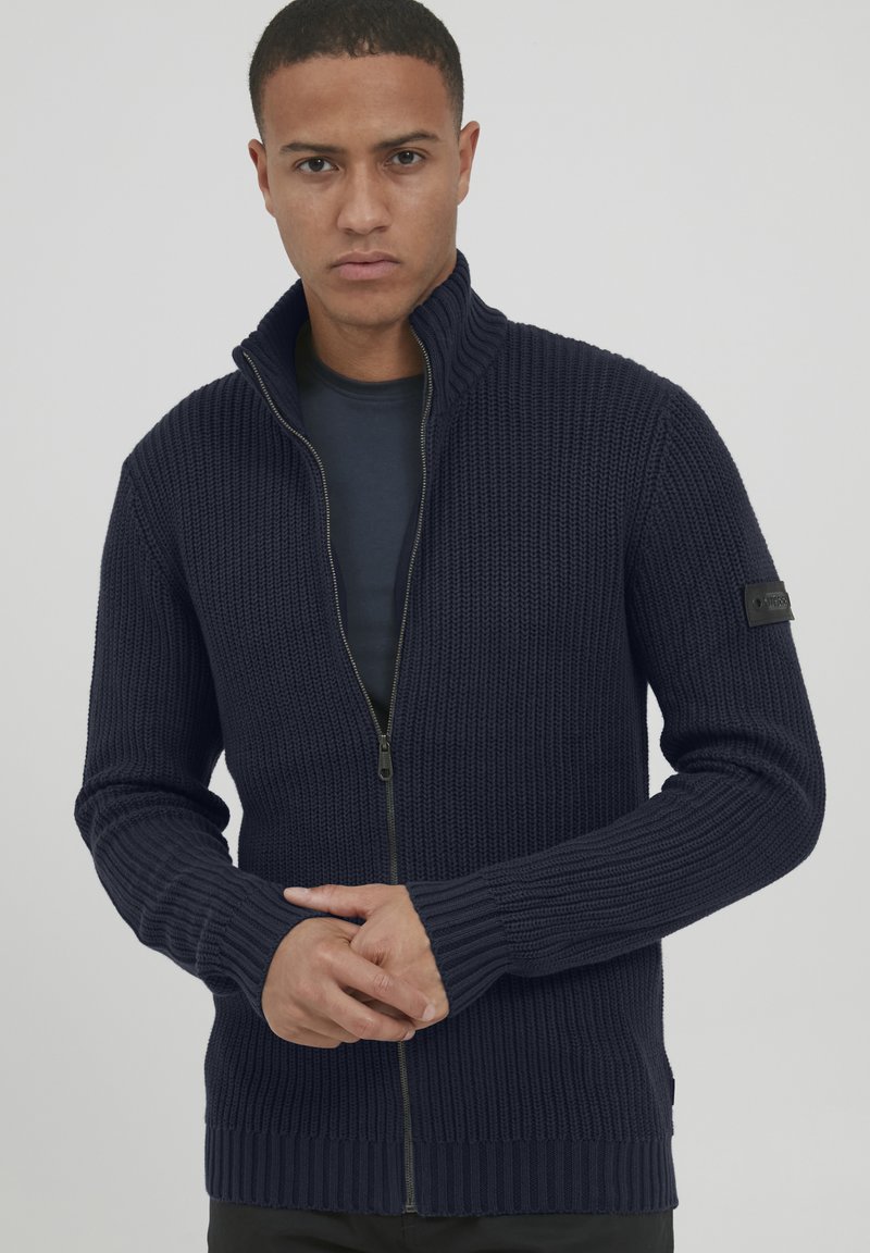 Maglione lavorato a maglia di un blu navy scuro con colletto alto, chiusura con zip, polsini a coste e un tessuto testurizzato. Presenta una patch con logo sul braccio.