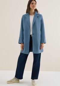 Abrigo de mezcla de lana de color azul claro con botones frontales y mangas anchas, combinado con una blusa blanca y pantalones de pierna ancha de color azul oscuro.