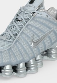 Ανοιχτό γκρι αθλητικό παπούτσι Nike Shox με διχτυωτό επάνω μέρος, κλείσιμο με κορδόνια, μεταλλική ασημί λογοτύπηση και χαρακτηριστική σόλα με ελατηριοειδή απορρόφηση κραδασμών.