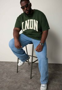 Grøn grafik t-shirt med "LNDN" tekst i creme, parret med lyseblå jeans og hvide sneakers. Person sidder på en grå skammel.