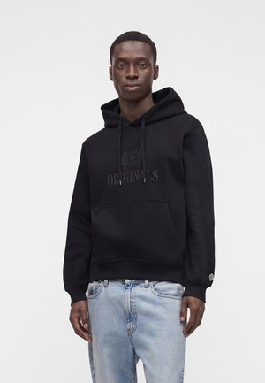 Homme portant un sweat à capuche noir avec le texte "Originals" et un jean bleu clair, debout devant un fond blanc uni.