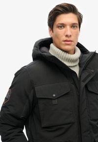 Sort parka-jakke med hætte, der har to brystlommer, en struktureret grå turtleneck under og et broderet logo på ærmet.