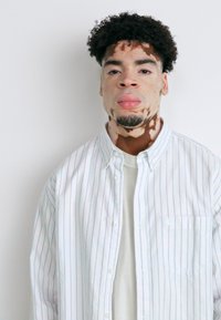 Witte, knoopsluiting shirt met blauwe verticale strepen, gedragen over een lichtgekleurde T-shirt. De man heeft kort, krullend haar en gezichtskenmerken.