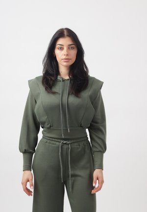 Vrouw met lang donker haar, gekleed in een olijfgroene hoodie met gewatteerde schouders en bijpassende broek met trekkoord, staand tegen een effen lichte achtergrond.