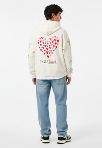 Felpa crema con una grafica a forma di cuore rosso e la scritta "SWEET heart" sul retro. Gli accenti includono piccoli disegni floreali sulle maniche. Abbinata a jeans azzurri chiari.