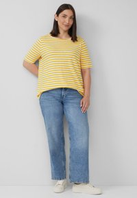 Gelb- und weiß gestreiftes T-Shirt mit kurzen Ärmeln, lockere Passform, kombiniert mit hellblauen weit geschnittenen Jeans und beigen Sneakers. Einfarbiger Hintergrund.