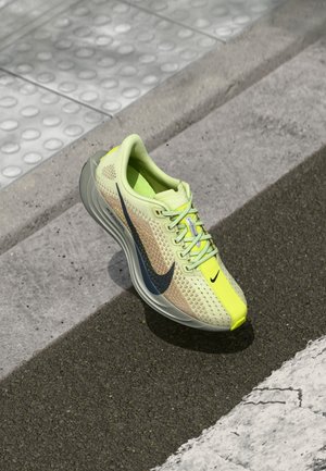 Lysegul og beige Nike løbesko med sort swoosh på fortov nær tekstureret hvid kant.