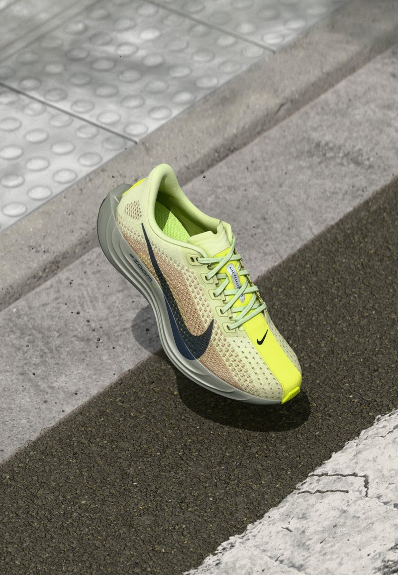 Chaussure de running Nike jaune clair et beige avec un swoosh noir sur le pavé près du bord de trottoir blanc texturé.