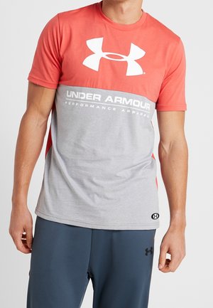 Mężczyzna w czerwono-szarej koszulce sportowej Under Armour i ciemnoniebieskich szortach na jednolitym jasnym tle.