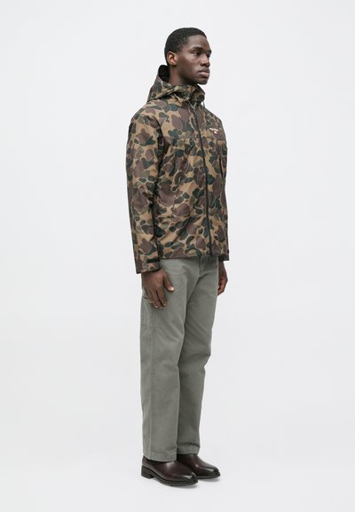 Vīrietis stāv, valkājot kapuci ar maskēšanās rakstu, pelēkas bikses un tumši brūnas zābakus, nedaudz pagriezies pa labi uz vienkārša fona.